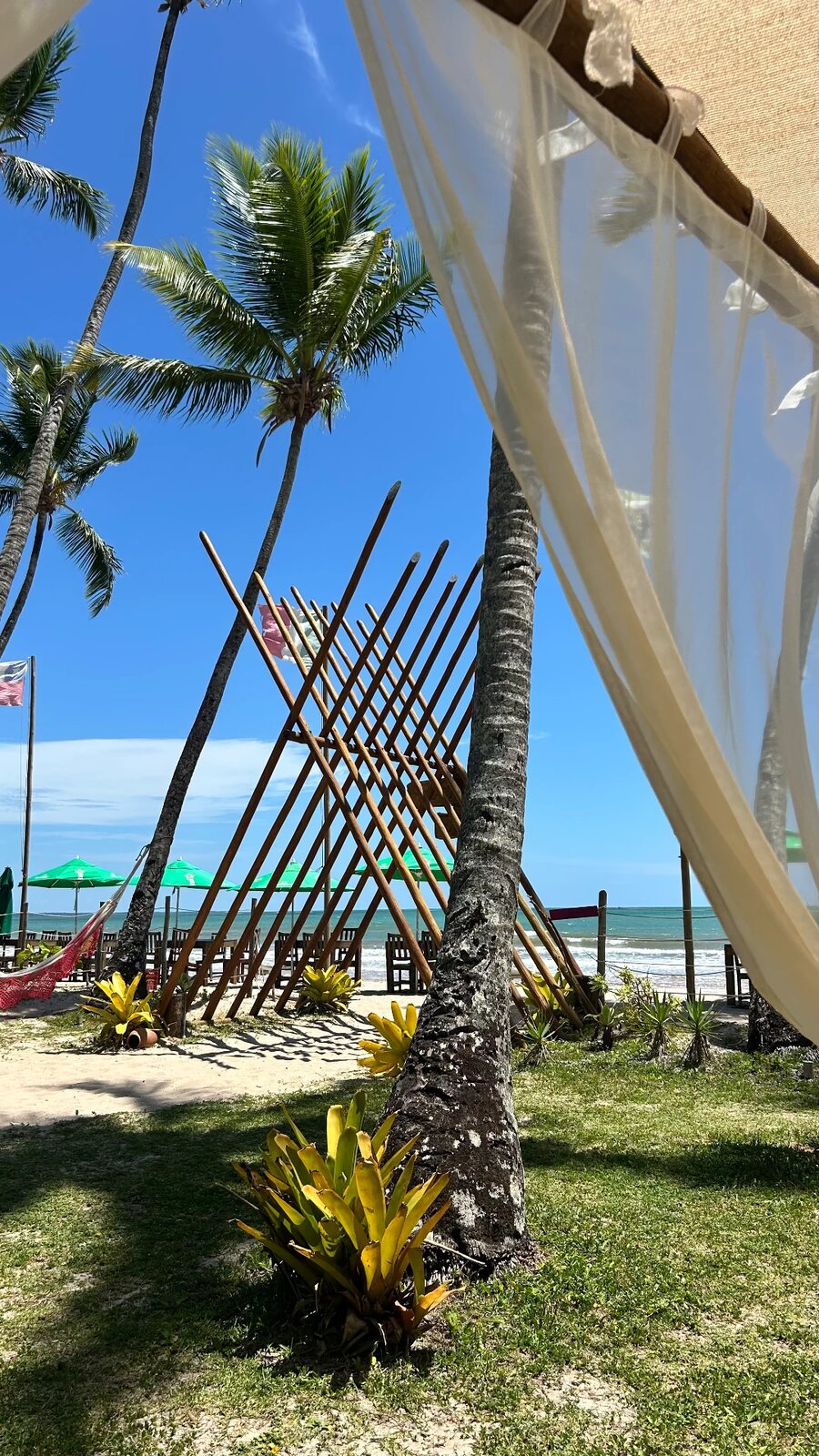 Entrada de restaurante de praia em Boipeba com véu branco esvoaçando entre coqueiros e o mar ao fundo